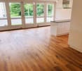 Empty room with light wooden flooring, white walls, large windows, and a small white kitchen unit in the corner.