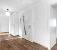 Bright, modern hallway with white walls, wooden flooring, and a built-in white cabinet. A coat rack is mounted on the wall, and a round ceiling light hangs above. The hallway leads to a bathroom with dark tiles visible in the background.