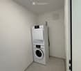 A small, empty room with light-colored tiled flooring and white walls. A white washing machine and dryer are stacked against one wall, and a door is visible on the right side.