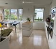 Open-plan living and dining area with a grey sofa, yellow chair, dining table, and modern kitchen island. Large windows offer a view of greenery.