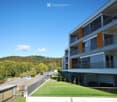 Modern multi-story apartment building with balconies and orange shutters, overlooking a green landscape and road.