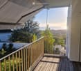 Balcony with wooden decking, metal railing, retractable awning, overlooking a scenic view of trees and the ocean at sunset.