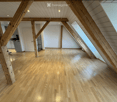 An empty attic space with wooden beams and exposed wooden floorboards, featuring large skylight windows and white walls.