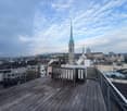 Wooden balcony with metal railing, overlooking a cityscape with historic buildings and a church spire.