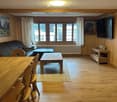 Cozy living and dining area with wooden walls, hardwood floor, large windows, sofa, coffee table, dining table, chairs, and TV stand.