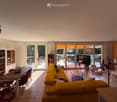 Bright living room with large windows and sliding glass doors leading to a patio. Features a yellow sectional sofa, wooden dining table with chairs, bookshelf, and a TV on a stand.