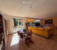 Bright living room with large glass doors leading to a patio, yellow sofa, wooden dining table with chairs, bookshelf, and television.