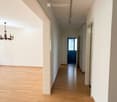 Empty hallway with wooden flooring, white walls, and a chandelier hanging from the ceiling. A window with a curtain is visible on the left side, and a blue door is at the end of the hallway.
