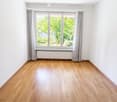 Empty room with light wooden flooring, white walls, large window with grey curtains, and a radiator below the window.
