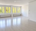 Empty room with light wooden flooring, white walls, large windows with white frames, and radiators beneath the windows.