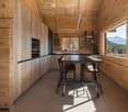 Modern kitchen with wooden walls and ceiling, large windows offering a mountain view, black countertop, and a small dining area with a table and stools.
