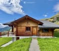 A wooden chalet with a sloped roof, stone foundation, and a small balcony overlooking a grassy area and mountain backdrop.