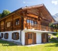 A two-story wooden house with a balcony, large windows, and a view of mountains in the background.