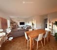 Bright living and dining area with wooden flooring, a grey sofa, a wooden dining table with white chairs, a television, and decorative shelves.
