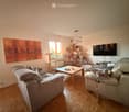 Bright living room with wooden flooring, a grey sectional sofa, a coffee table, a cat tree, a bookshelf, and a television mounted on the wall.