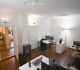 Bright, modern office space with multiple desks, chairs, laptops, monitors, and a small fireplace. The room features wooden flooring and white walls.