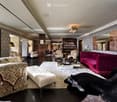 Elegant living room with patterned wallpaper, dark hardwood floors, and a mix of plush seating including a red velvet sofa, floral-patterned armchairs, and a white ottoman. A coffee table sits in the center, flanked by additional seating and a large mirro