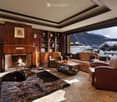 Elegant living room with a fireplace, leather sofa, armchairs, coffee table, bookshelf, and a scenic mountain view through large windows.