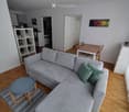 Modern living room with a grey sectional sofa, wooden floor, small round stool, dining table with chairs, white shelving unit with decorative items, and a potted plant.