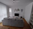 Modern living room with a grey sectional sofa, coffee table, TV stand, white shelving unit, and floor lamp. Large windows with white blinds and a view of the city.