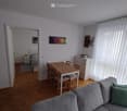 Bright, open-plan living and dining area with a grey sofa, wooden dining table with chairs, a small bedroom visible through an open doorway, and a nightstand beside the bed.