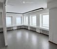 Empty room with white walls, grey carpeted floor, multiple windows with blinds, radiators, and fluorescent lighting.