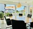 Bright dining room with a large round wooden table surrounded by black chairs, a potted plant on the left, and large glass doors leading to a patio with outdoor furniture and an umbrella.