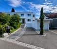 Modern two-story house with white exterior, multiple windows, garage, and landscaped garden with trees and shrubs.
