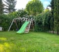 Lush garden with a green slide, swing, and climbing frame surrounded by trees and flowering bushes.