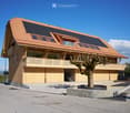 Modern two-story house with a wooden facade, solar panels on the roof, large windows, and a balcony overlooking the surrounding landscape.