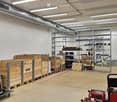 Empty warehouse space with concrete floor, exposed brick wall, metal shelving units, and storage boxes.