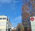 Modern multi-story building with signage for "Progile" and "Riedwiesen23" visible, surrounded by trees and parked cars.
