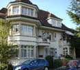 Two-story house with white exterior, multiple windows, and a balcony on the upper floor. A blue car is parked in front of the house.