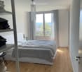 A modern bedroom with a wooden floor, a large bed with grey bedding, a chandelier, a shelf with folded clothes, and a window with curtains offering a scenic view.