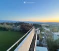 A rooftop terrace with a grassy area, metal railing, and a panoramic view of a cityscape and mountains at sunset.