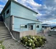 Modern two-story building with light blue exterior, concrete steps leading to entrance, small garden area with shrubs and rocks, and a view of the landscape.