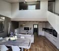 Modern open-plan living and dining area with a white sofa, wooden dining table, white chairs, and a kitchen area with white cabinets and stainless steel appliances.