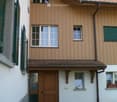 Two-story house with wooden siding, multiple windows, and a tiled roof. Features a wooden front door with glass panels, a small garden area, and decorative items on the windowsills.