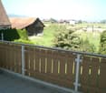 Balcony with wooden railing overlooking a green lawn, trees, and distant houses.