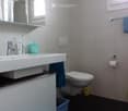Modern bathroom with white tiles, a large sink with a mirror, a toilet, and a blue towel hanging on the wall. A small blue rug is placed on the dark floor.