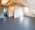 Empty attic room with exposed wooden beams, grey walls, dark grey floor, and two windows allowing natural light.