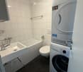 Modern bathroom with white tiled walls, black tiled floor, sink, bathtub, toilet, and stacked washing machine and dryer.