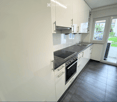 Modern kitchen with white cabinets, dark countertops, induction cooktop, oven, and sink. Large window offers a view of the garden.