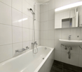Modern bathroom with white tiled walls and floor, featuring a bathtub with a handheld shower, a sink with a modern faucet, and a large mirror with built-in lighting.