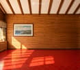 Empty room with wooden walls, red tiled floor, large window with view, small table with plants and lamp, framed picture on wall.