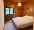 Cozy wooden-paneled bedroom with a double bed, wooden chair, radiator, and window with curtains.