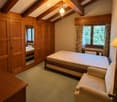 Cozy attic bedroom with wooden beams, a large bed, a sofa, a wardrobe, and a mirror. The room features exposed wooden beams on the ceiling and warm wooden paneling on the walls.