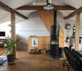 A spacious living room with wooden beams, a central black fireplace, a sofa, a chair, a small table, a screen, and a potted plant.