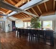 A spacious, modern room with wooden flooring and exposed wooden beams on the ceiling. A large central table is surrounded by several high-backed chairs. An arcade machine is positioned against the wall, and a chessboard is set up on the table. A large gre