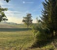 A peaceful rural landscape with a grassy field, trees, and a distant view of cultivated farmland under a clear blue sky.